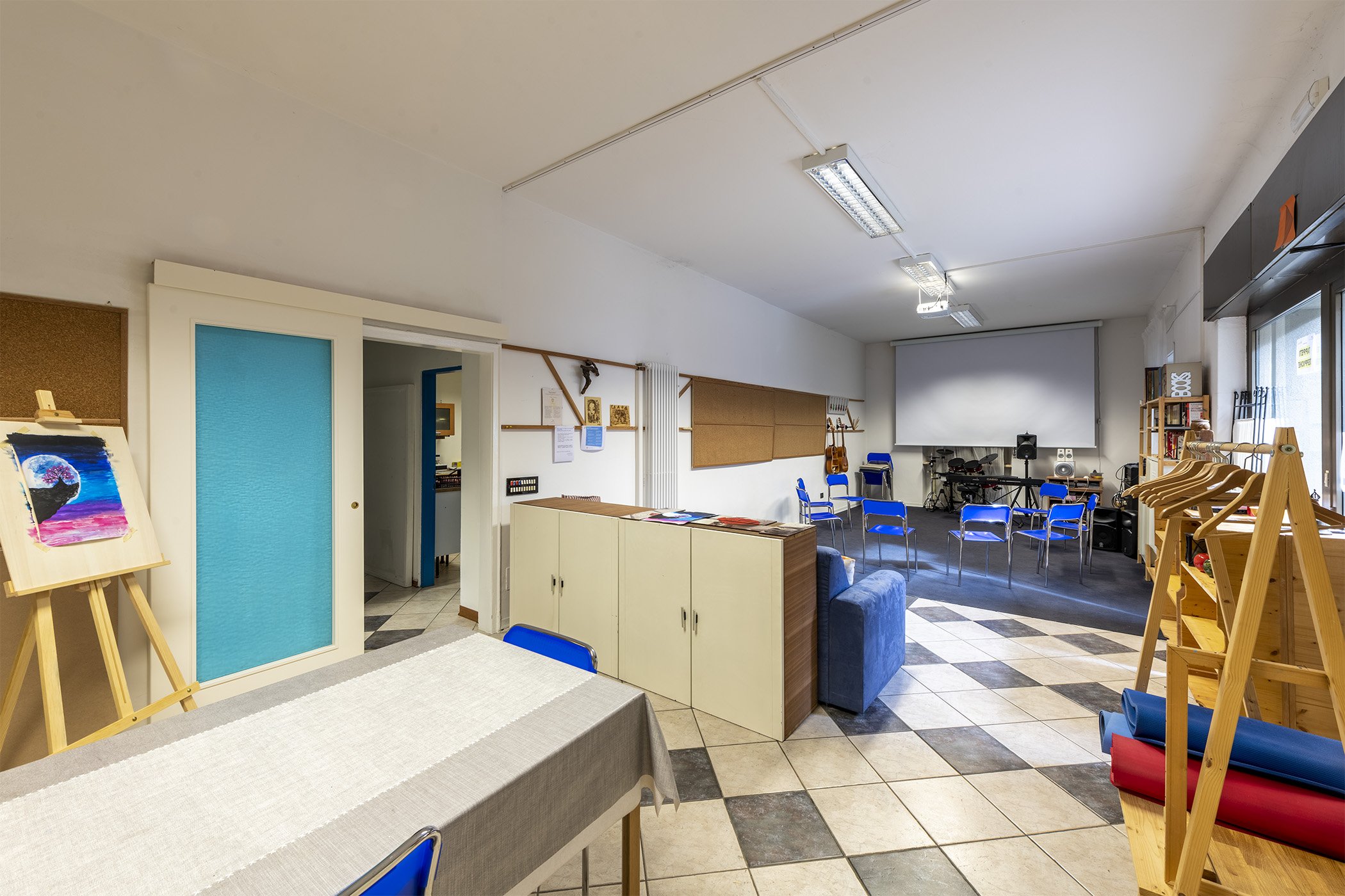 Sala polifunzionale con sedie disposte a semicerchio, schermo per proiezioni, strumenti musicali sul fondo, cavalletto con dipinto e area laboratorio con tavolo e scaffali in legno
