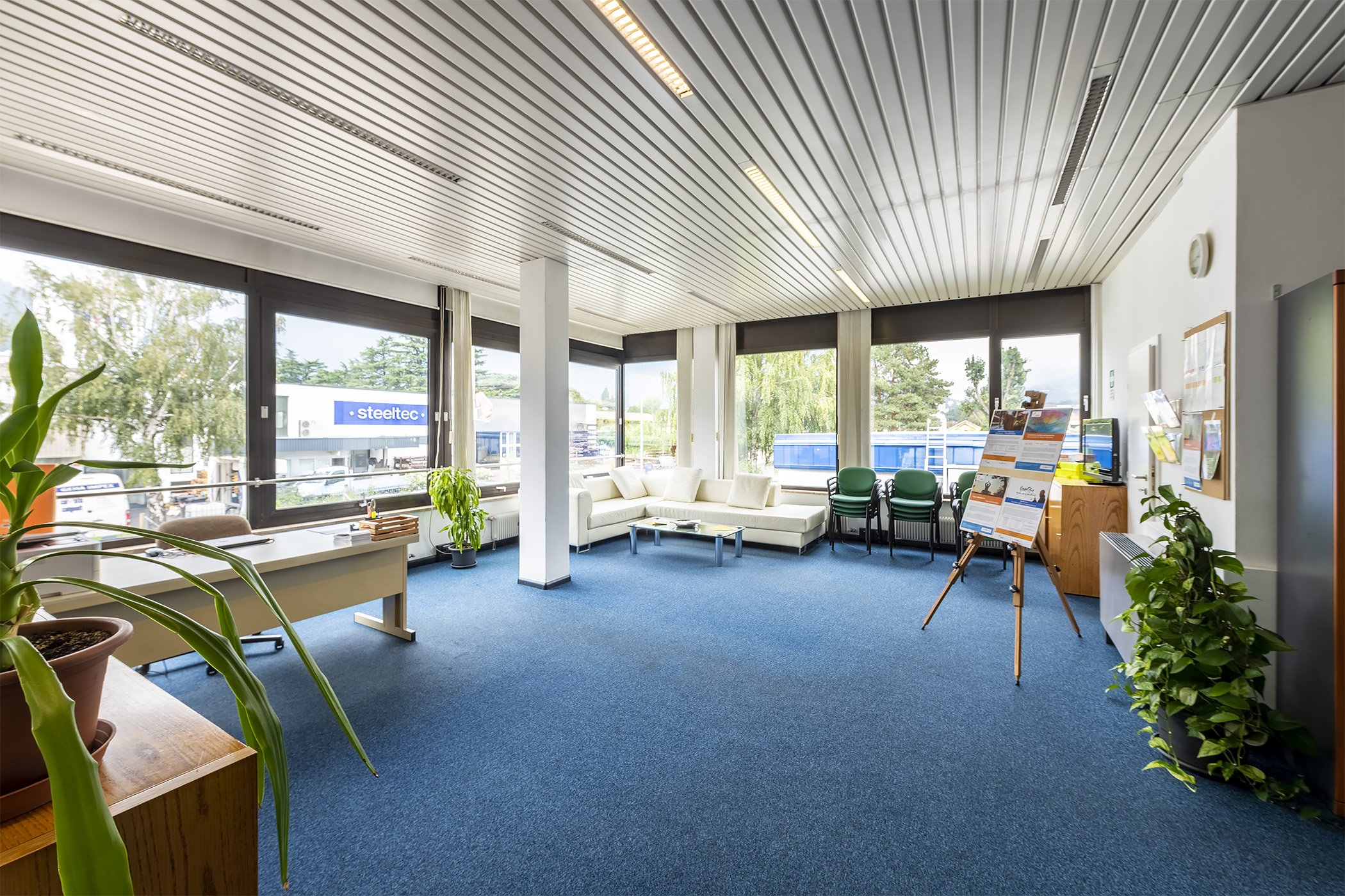 Sala luminosa con ampie vetrate, divano bianco, sedie impilate, scrivania, piante e cavalletto con pannello informativo